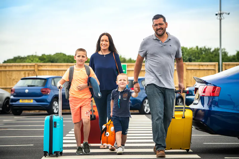 Family with luggage crossing pedestrian path in Gatwick Airport parking lot by blue cars.