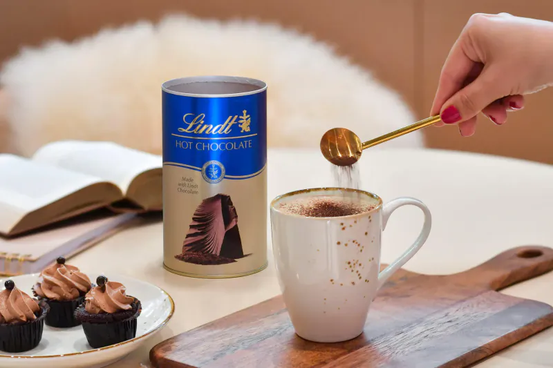 Hand stirring hot chocolate in white mug with Lindt Hot Chocolate can, cupcakes on wooden board nearby.