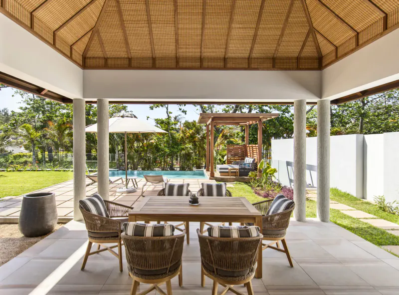 Open-air pavilion with wooden dining table and chairs overlooking tropical garden and infinity pool at Anantara Iko Mauritius resort.