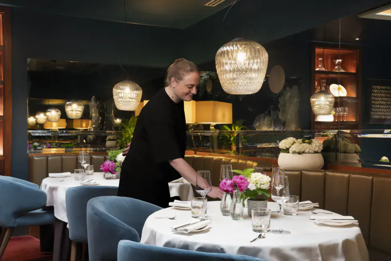 Server in black uniform arranging pink flowers on white tablecloth at elegant restaurant with blue chairs and pendant lamps