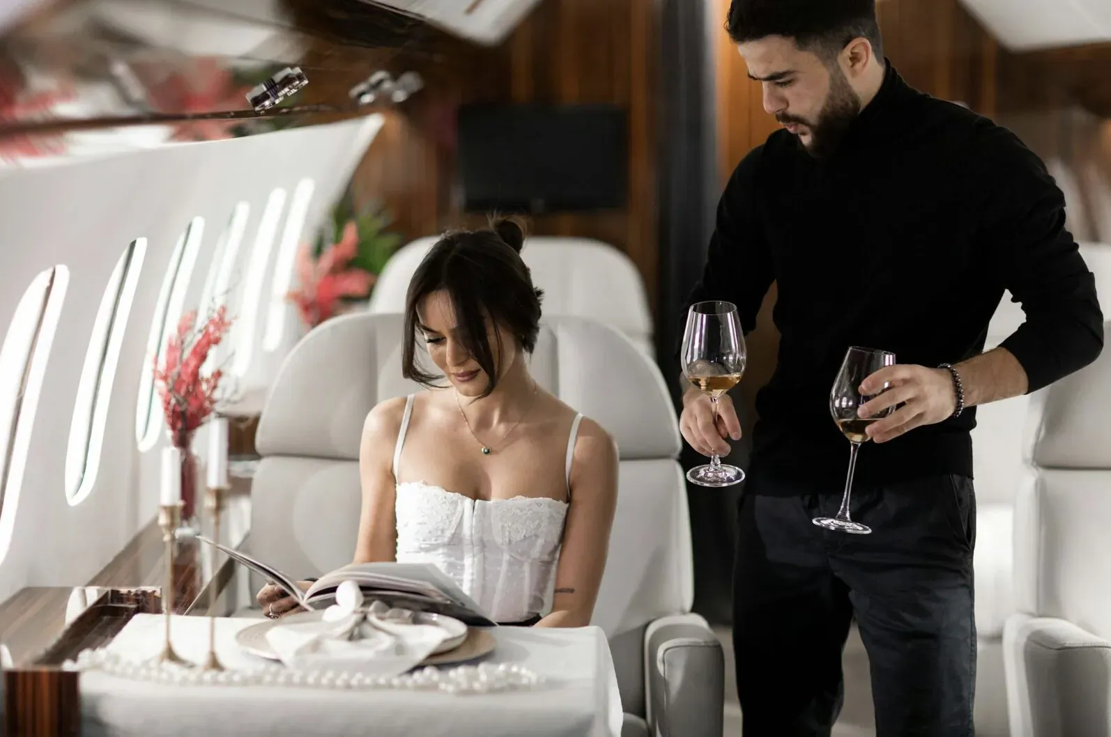 Elegant couple in private jet: man in black offers wine to woman in white dress at candlelit table with flowers.