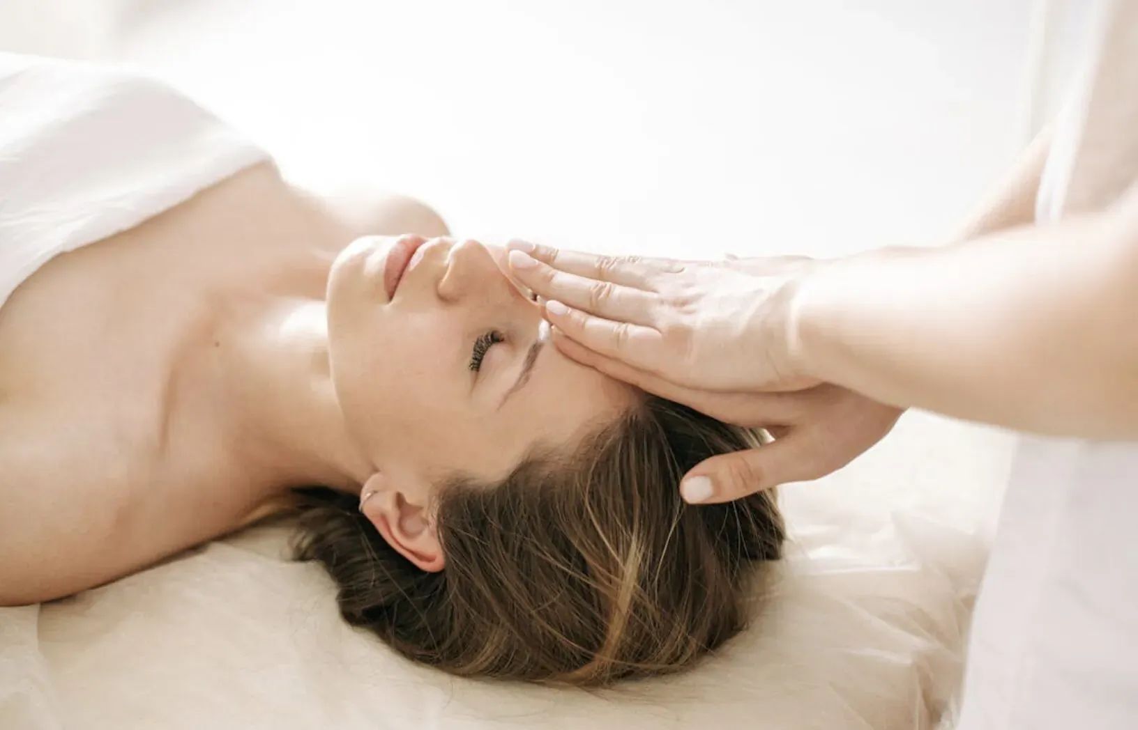 Holistic therapist giving head massage to relaxed woman lying on table, wrapped in white towel