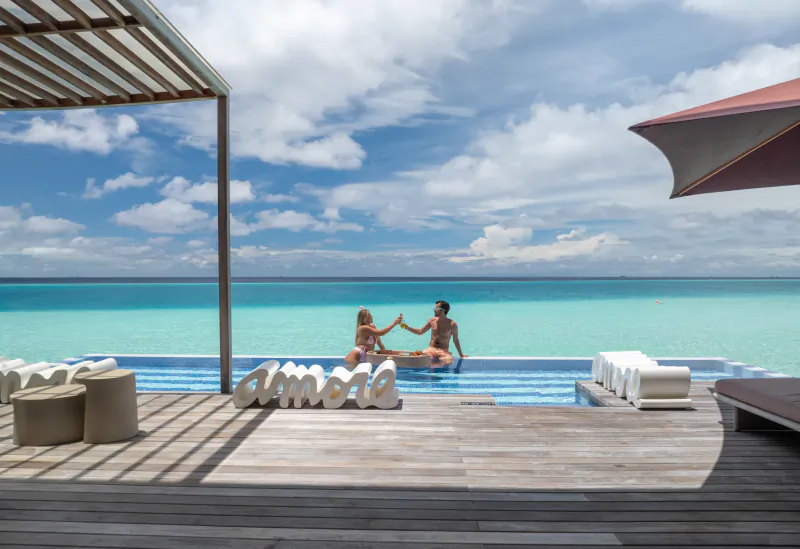 Couple high-fiving in pool at Crossroads Maldives resort, turquoise lagoon, 'AMOR' sign, shaded deck, sunny sky