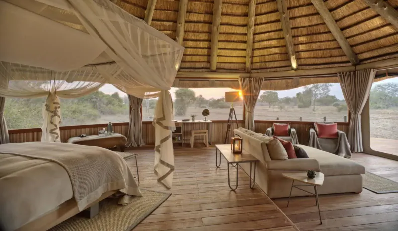 Luxurious thatched safari tent interior with king canopy bed, sofa, desk, and sunset savanna views at Lion Camp.