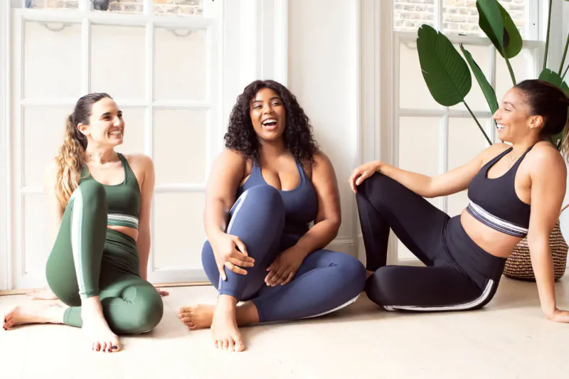 Three diverse women laughing joyfully on floor in colorful leggings and sports bras by window with plants
