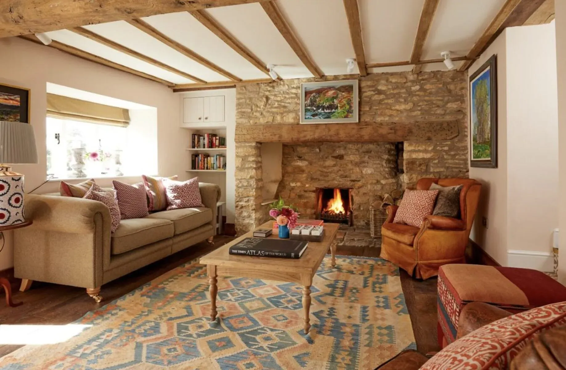 Cozy Cotswolds inn living room with stone fireplace lit, beige sofa, armchair, colorful rug, and art on walls.