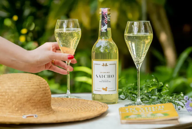 Hand holding flute of Saicho sparkling tea, bottle and another glass on table with straw hat amid lush greenery