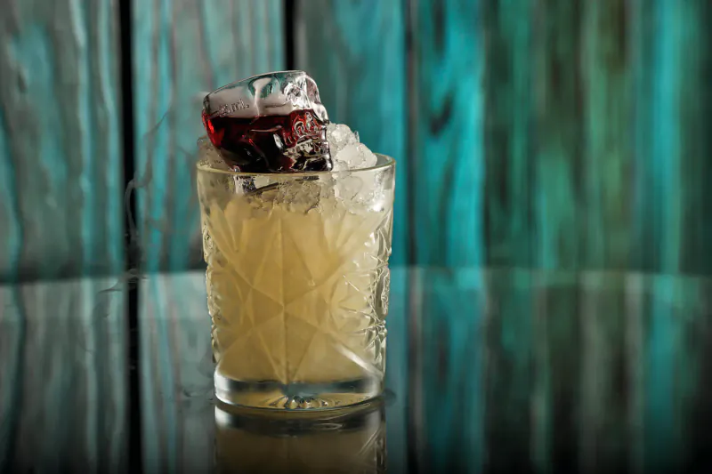 Close-up of etched highball glass with pale yellow cocktail, red ice cube, on reflective table against teal wood wall.