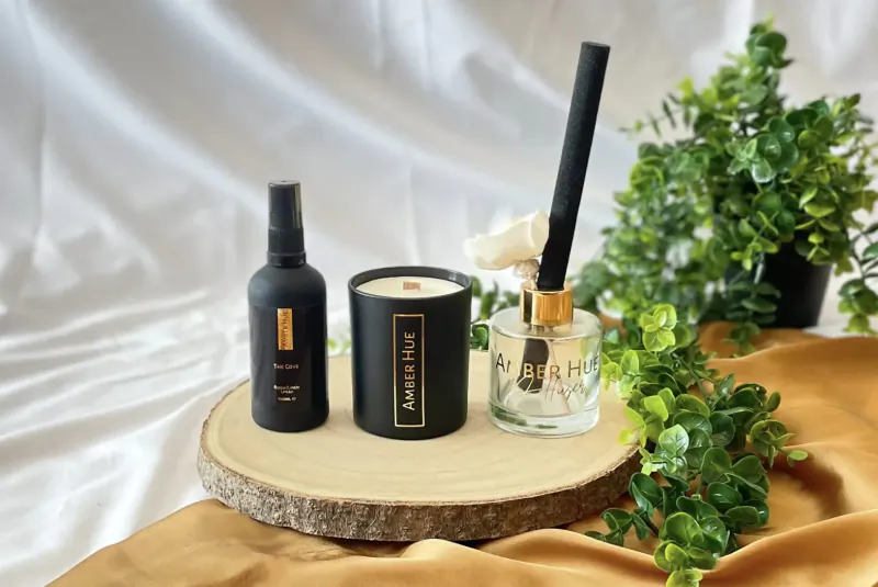 Amber Hue luxury black candles, spray, and reed diffuser on wood slice with green plants and gold backdrop