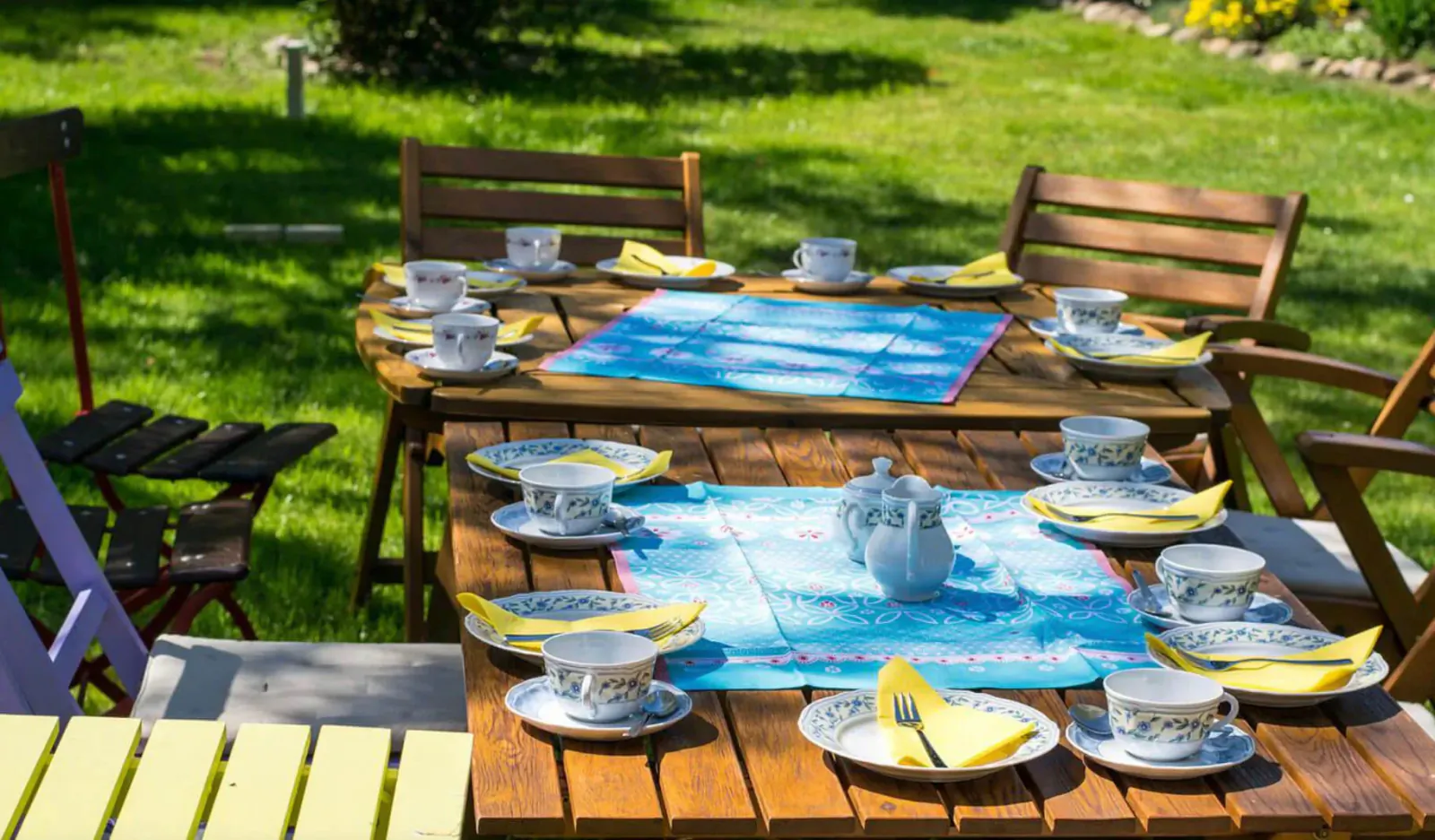 Outdoor wooden table set for al fresco tea with white cups, blue and yellow napkins, teapot on green lawn