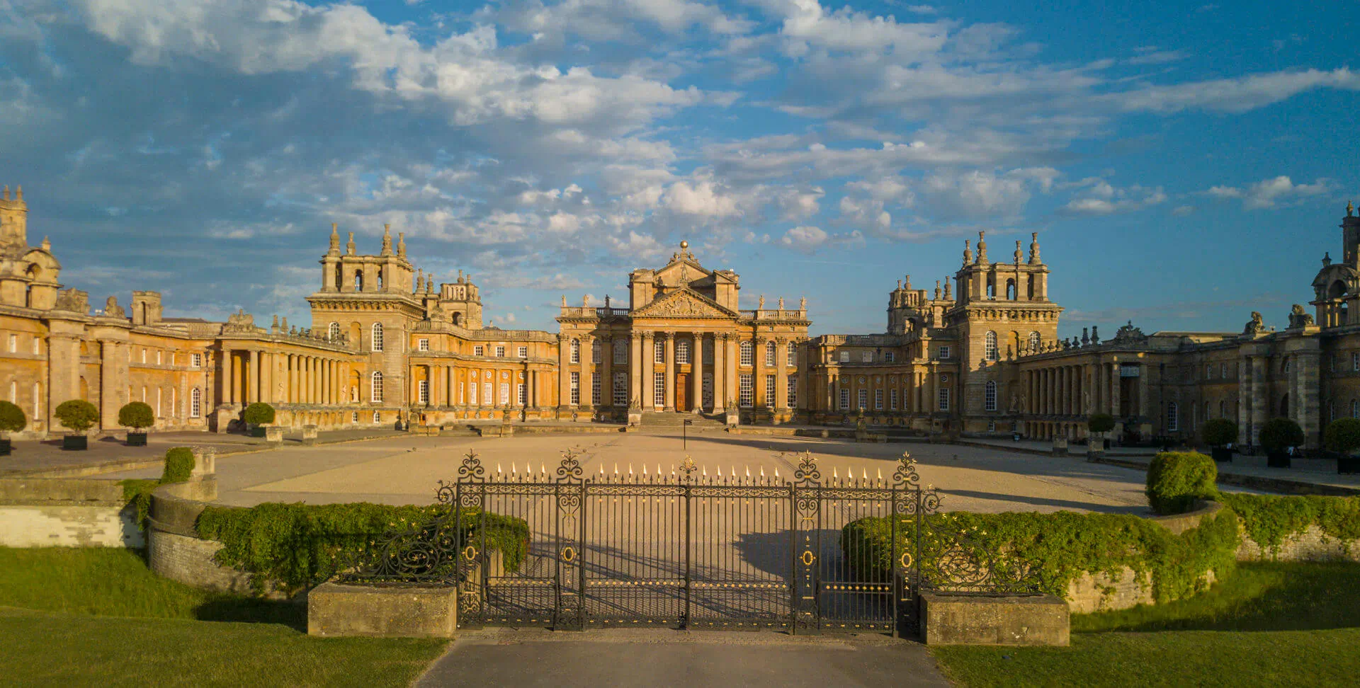 Aerial view of Blenheim Palace's grand golden facade and courtyard with ornate gates under blue sky.