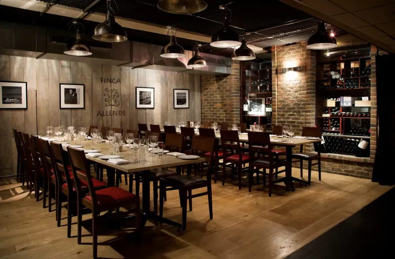 Elegant private dining room with long wooden table set for meal, red chairs, brick walls, wine racks, and pendant lights.