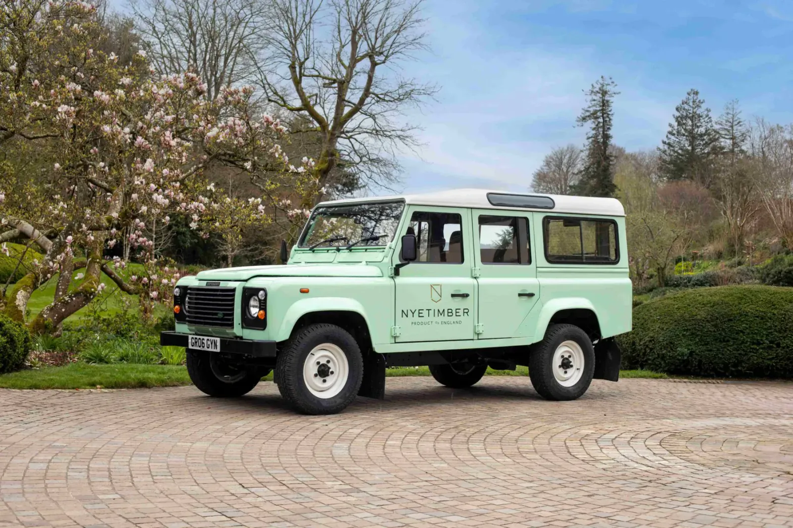 Pale green Land Rover Defender branded 'Nyetimber' parked on gravel amid spring cherry blossoms and trees under blue sky