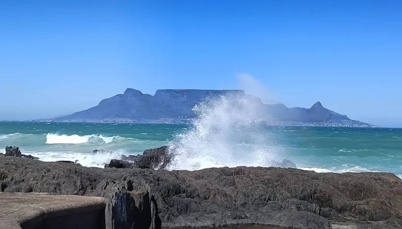 Table Mountain with cableway plume in background, crashing waves on rocky Cape Town coastline under blue sky