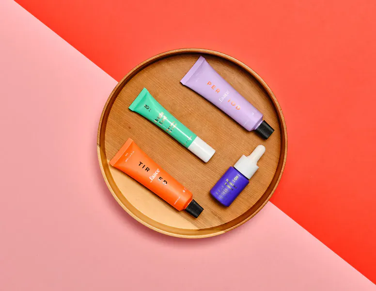 Wooden tray on pink-red gradient holding PFR skincare: orange, green, purple tubes and blue dropper bottle
