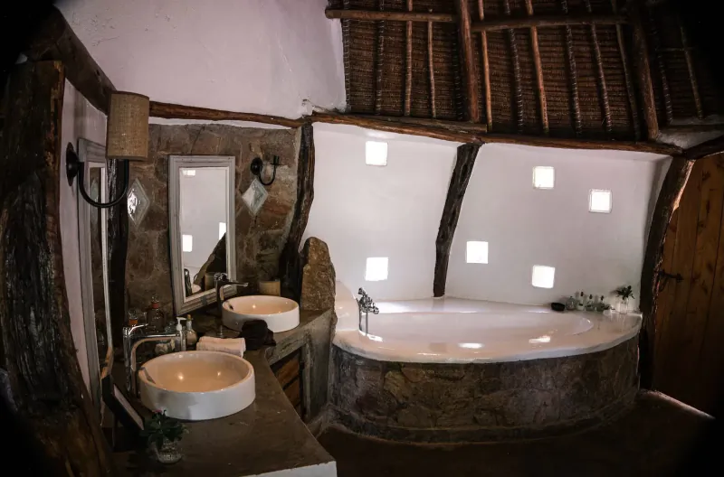 Rustic Kenyan bathroom in House In The Wild with stone double sinks, white bathtub, thatched ceiling, and wooden beams.