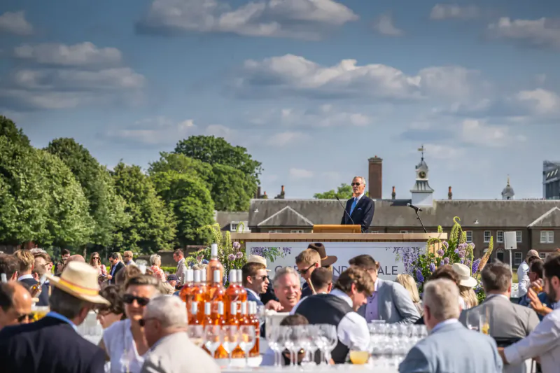 Elegant crowd at GOFFS London Sale x Château Léoube event, man speaking at podium, enjoying wine amid trees and buildings