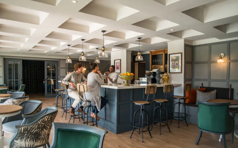 Three people chatting at a stylish wooden bar counter in Stanley & Livingstone Boutique Hotel lounge, with pendant lights and lemon bowl.
