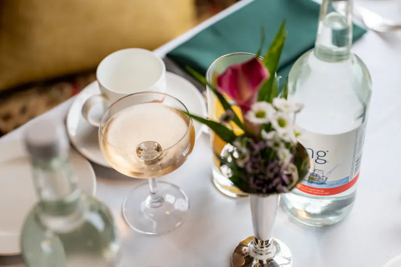 Table setting with rosé champagne, water bottle labeled 'Steam', tulips and daisies in silver vase, teacup on saucer
