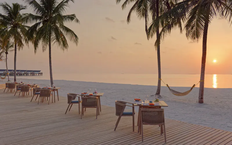 Sunrise over white sand beach at Maldives resort, wooden deck with wicker tables set for dining, palm trees and hammock.