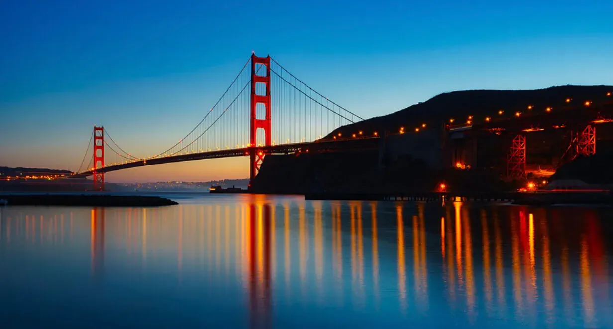Golden Gate Bridge at dusk, illuminated with lights reflecting on calm bay waters