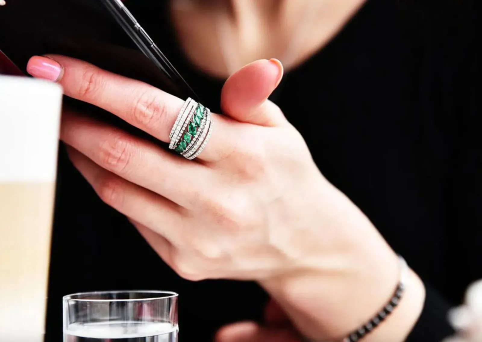 Woman's hand holding smartphone, showcasing stacked diamond and green gemstone ring, bracelet, near water glass
