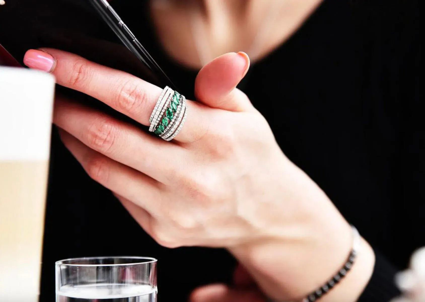 Woman's hand holding smartphone, showcasing stacked diamond and green gemstone ring, bracelet, near water glass