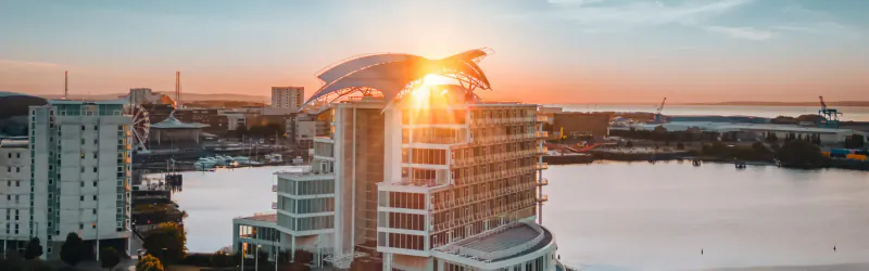Aerial view of modern white high-rise hotel with sail-like roof glowing in sunset over marina and cityscape.