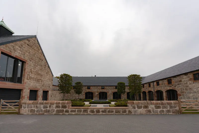 Ardross Whisky Distillery's stone barn-style buildings with slate roofs, manicured trees, and courtyard under cloudy sky