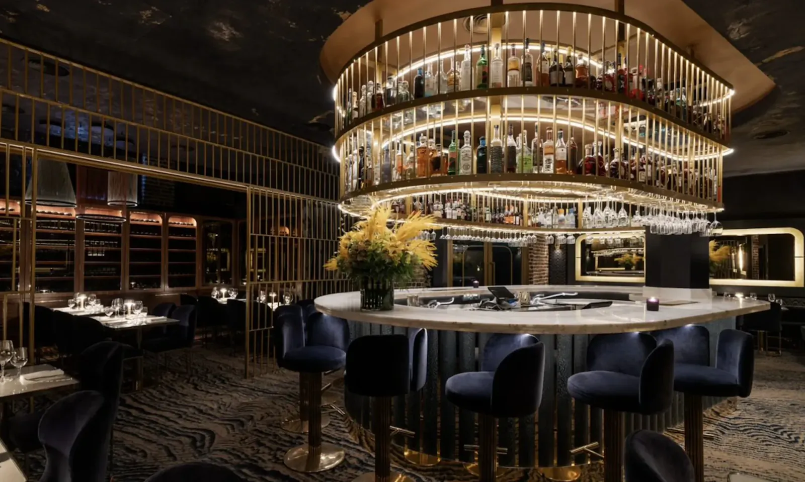 Elegant circular bar at Leven Manchester with tiered liquor shelves, yellow flowers, blue velvet stools, and tables.