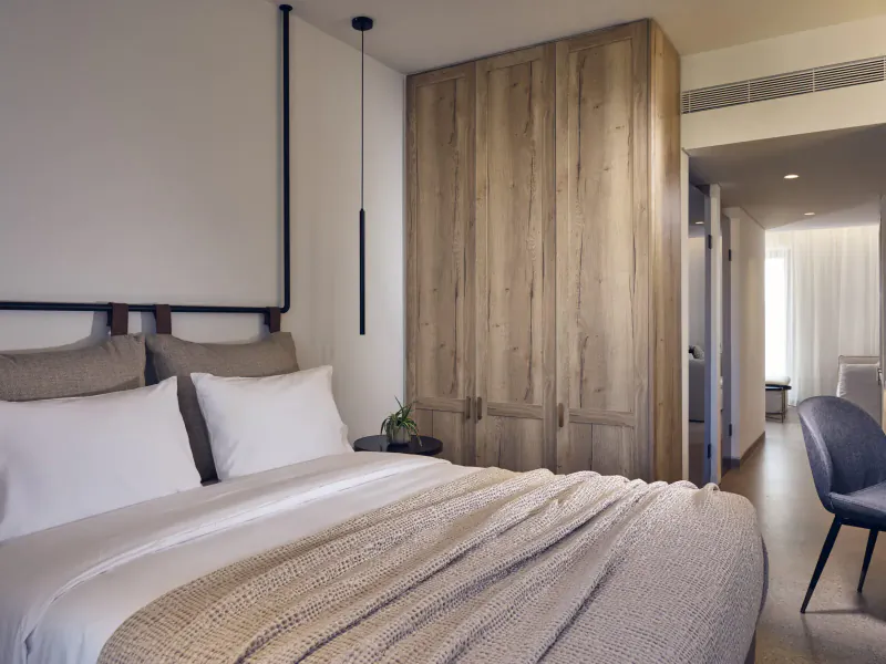 Modern hotel bedroom with king bed, beige throw, wood wardrobe, potted plant, and blue accent chair