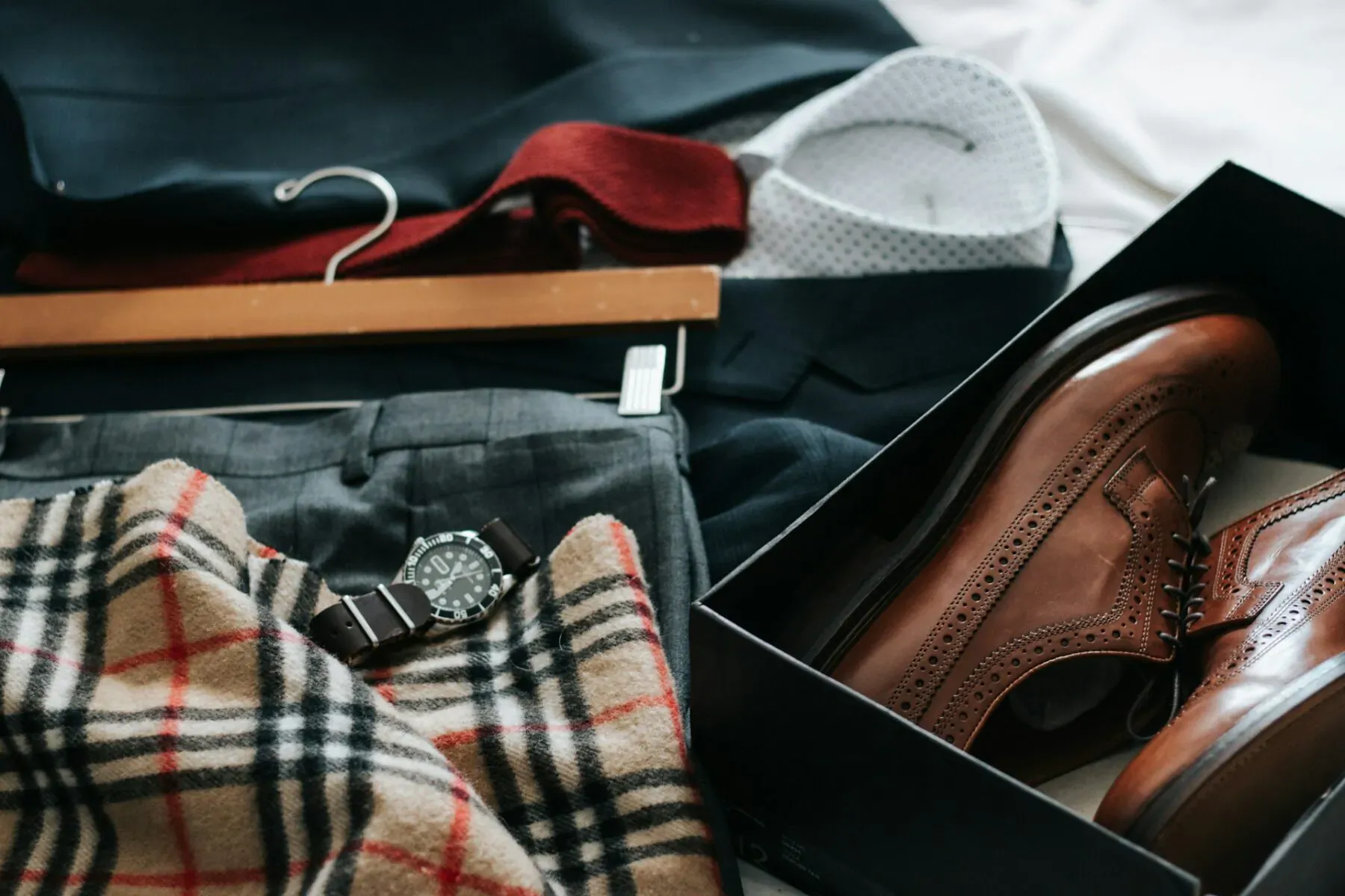 Men's wardrobe essentials: navy suit, trousers, red tie on hanger, white shirt collar, Burberry scarf, brown brogues in box.