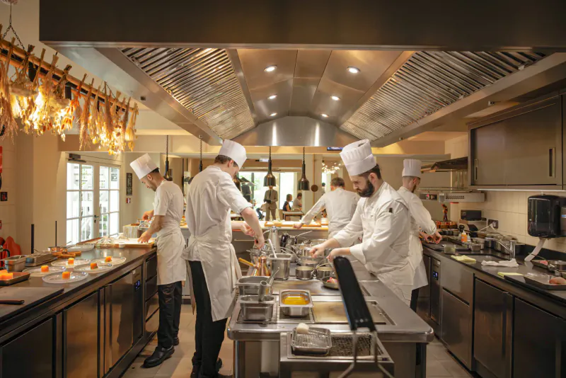 Busy professional kitchen with chefs in white hats and uniforms preparing dishes on stainless steel counters