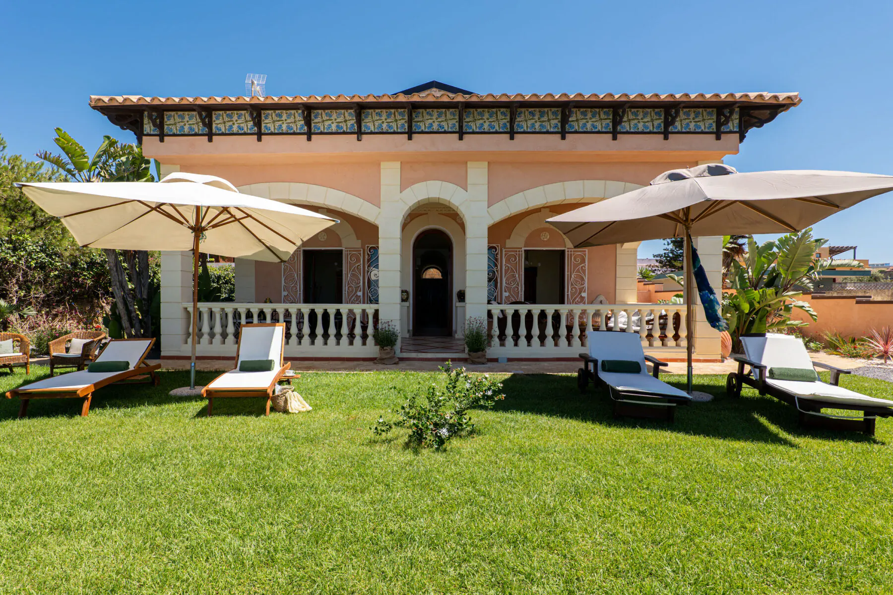 Peach-colored Mediterranean villa with arched porch, lounge chairs, umbrellas, and lush green lawn under blue sky