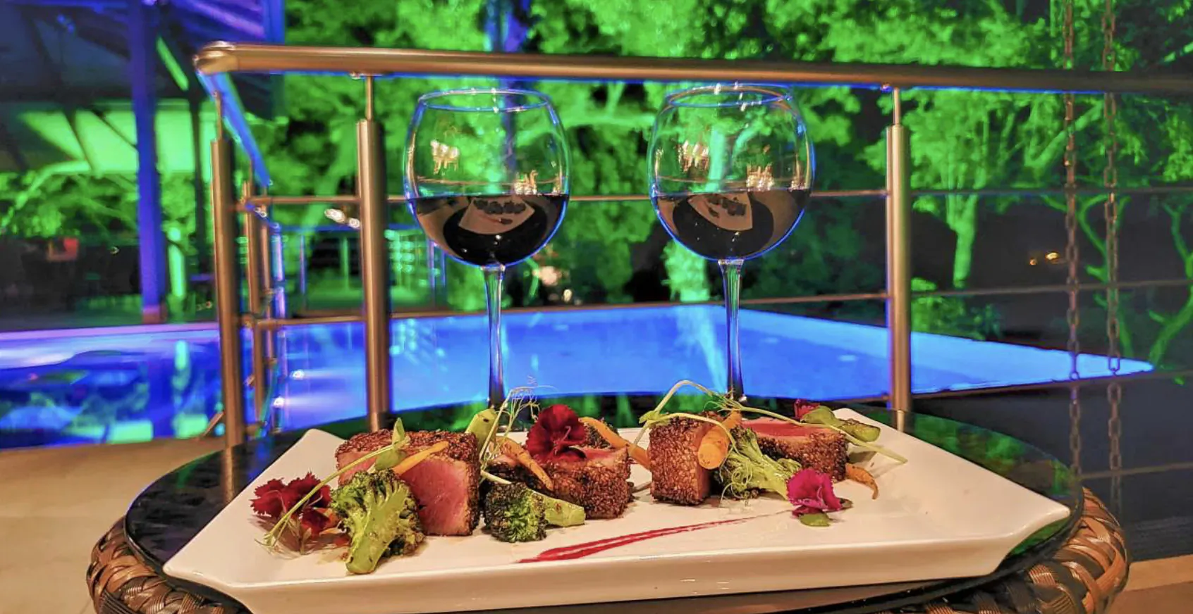 Luxury plated seared tuna with greens and sauce, flanked by red wine glasses, by infinity pool railing at night