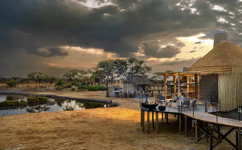 Camp Kala luxury thatched lodges on wooden deck by lagoon at Etosha sunset under stormy skies