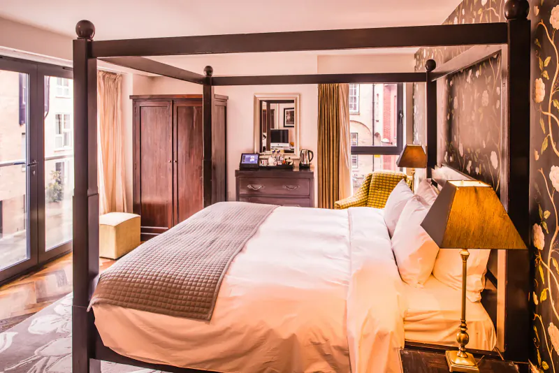 Elegant hotel bedroom with four-poster bed, wardrobe, lamps, and city view windows.