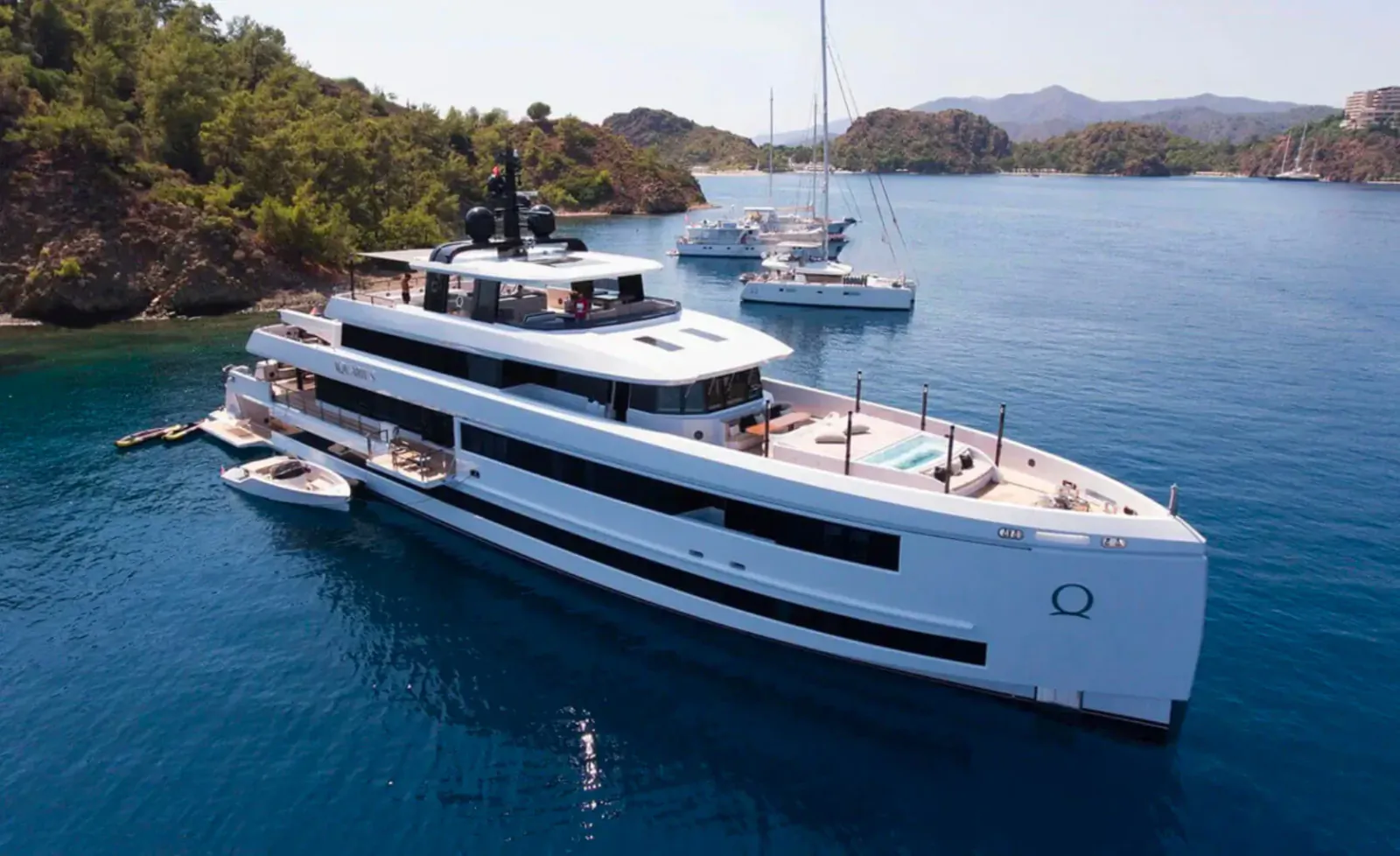 White luxury superyacht docked at a scenic Turkish bay with turquoise waters, green hills, and other yachts nearby.