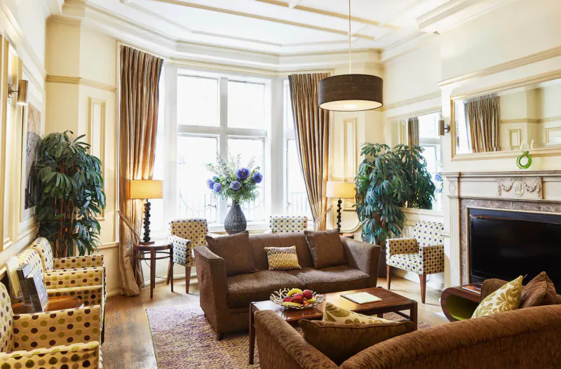 Elegant hotel lounge with bay windows, plush brown sofas, armchairs, plants, flowers, and fireplace.