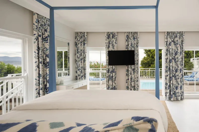 Spacious bedroom at The Plettenberg with four-poster bed, blue floral curtains, pool view, and mountain backdrop.