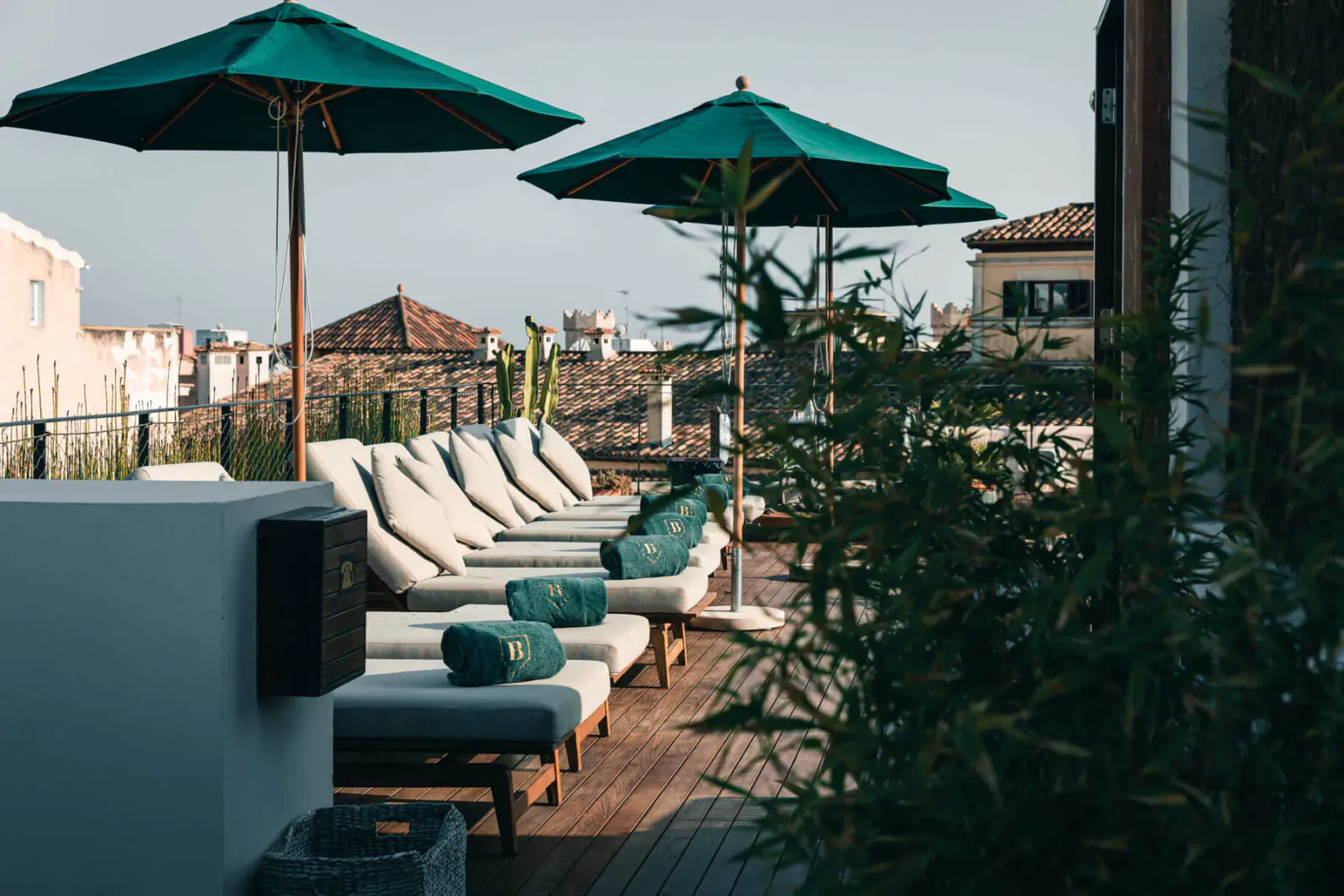 Luxury rooftop terrace with green umbrellas, cushioned loungers, potted palms, and terracotta rooftops in background