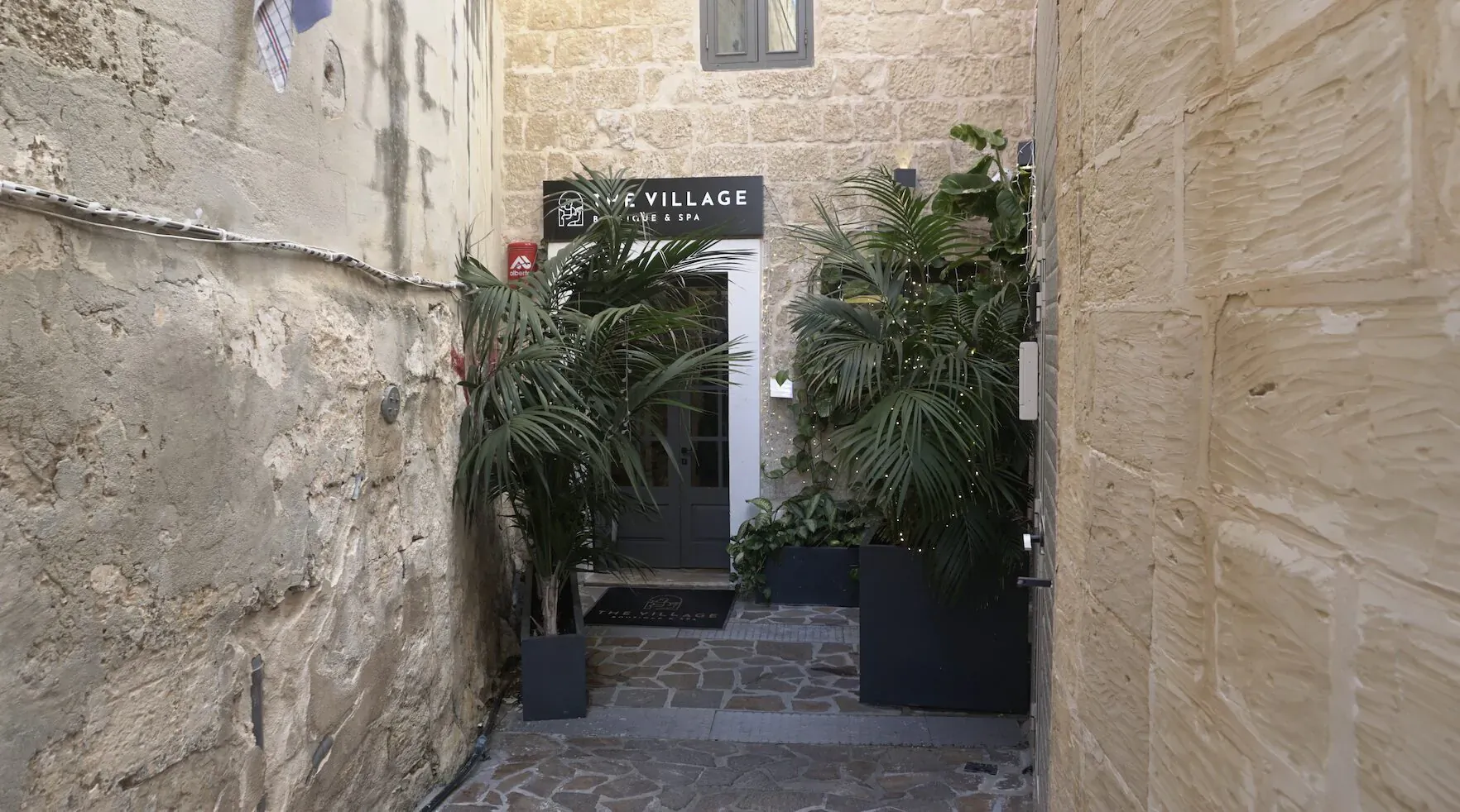 Narrow alley entrance to The Village flanked by palms and plants, white door, stone walls in Malta.