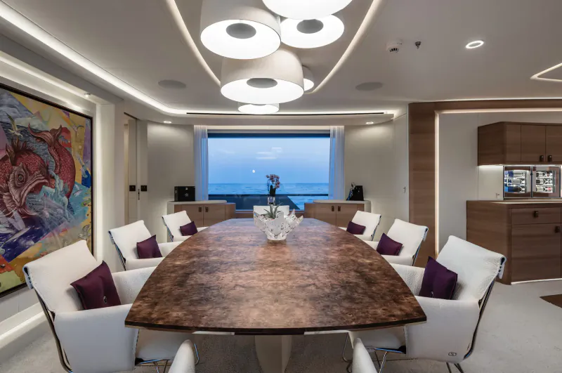 Luxurious yacht dining room with long brown table, white chairs, pendant lights, abstract art, and ocean sunset view