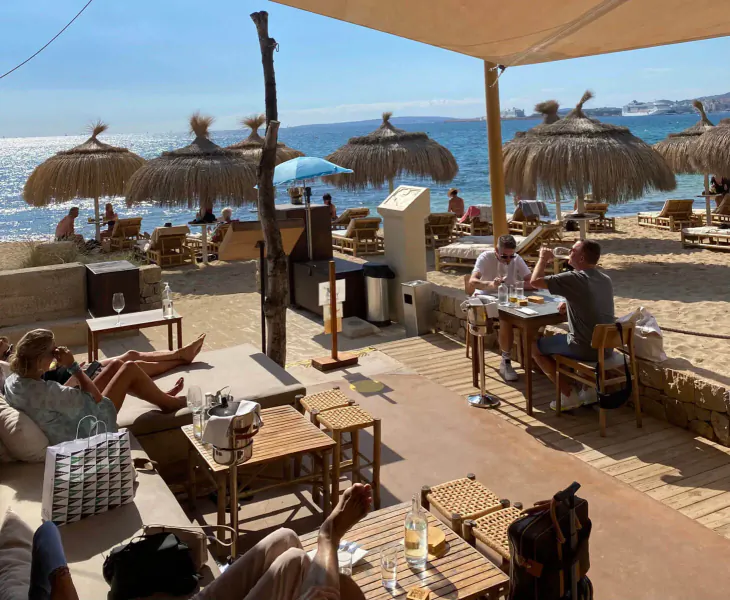 Assaona Gastrobeach Palma Club: patrons dining and relaxing on sunny beachfront terrace with thatched umbrellas and sea view.