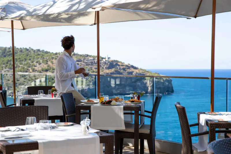 Man in white shirt stands at cliffside table with breakfast, overlooking sea at Jumeirah Port Soller Hotel terrace.