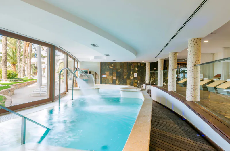 Luxury indoor pool with cascading waterfall, palm trees outside, lounge chairs, at Nixe Palace resort.