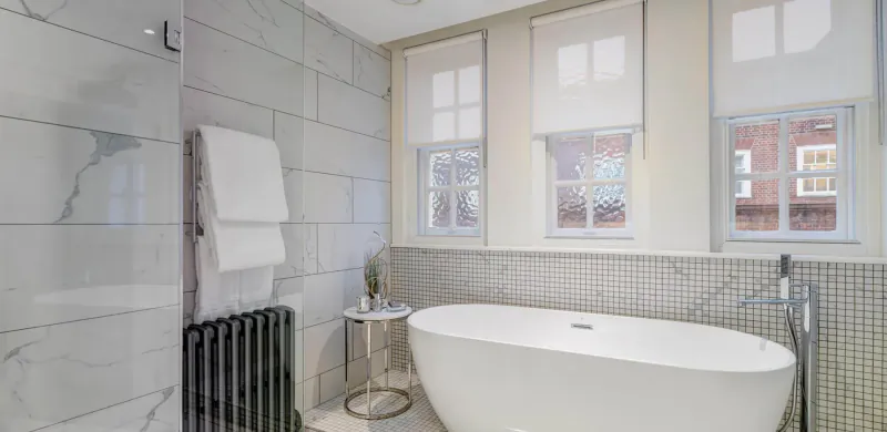Elegant modern bathroom at Apex Temple Court Hotel with freestanding white tub, gray tiles, towel radiator, and shuttered windows.