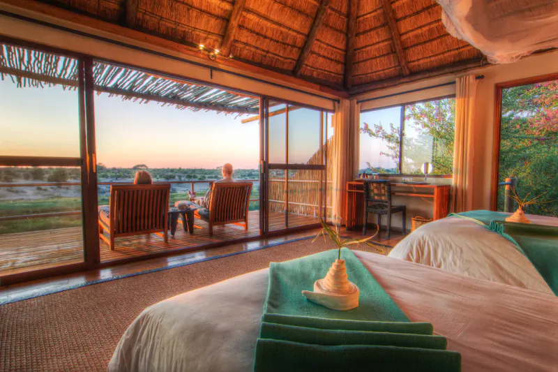 Luxurious thatched Tau Game Lodge suite with king bed, chairs on deck overlooking sunset savanna.