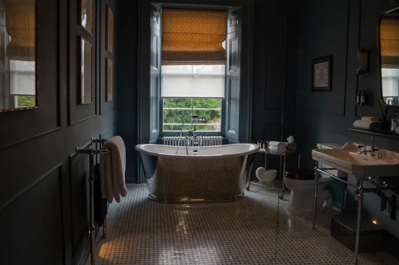 Elegant dark blue bathroom with freestanding bathtub, towels on heated rack, window with blinds, and tiled floor.
