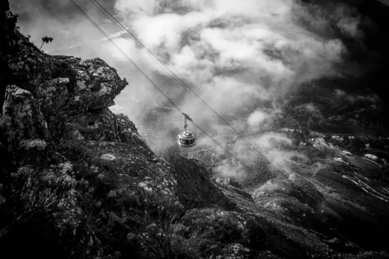 Black-and-white photo of Cape Town Aerial Cableway gondola soaring above misty cliffs and clouds on Table Mountain.